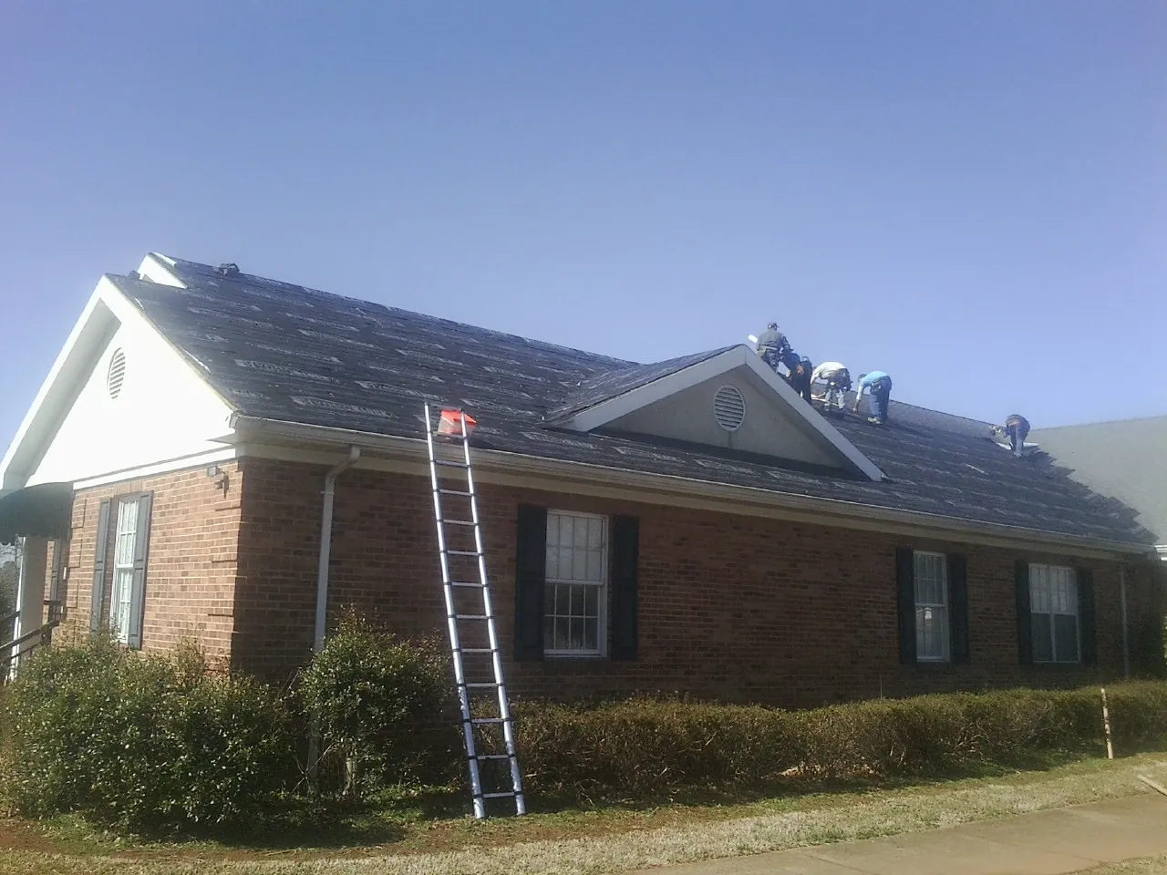 Crew tearing shingles off a brick ranch roof with a ladder set — Metal Roof Repair work by our crew in Bessie