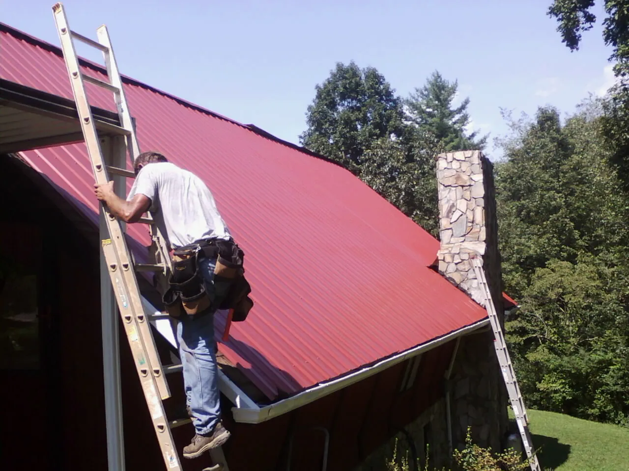 Roofer on a ladder beside a red metal roof and stone chimney — Soffit & Fascia Repair by Roofing Brothers in Bessie